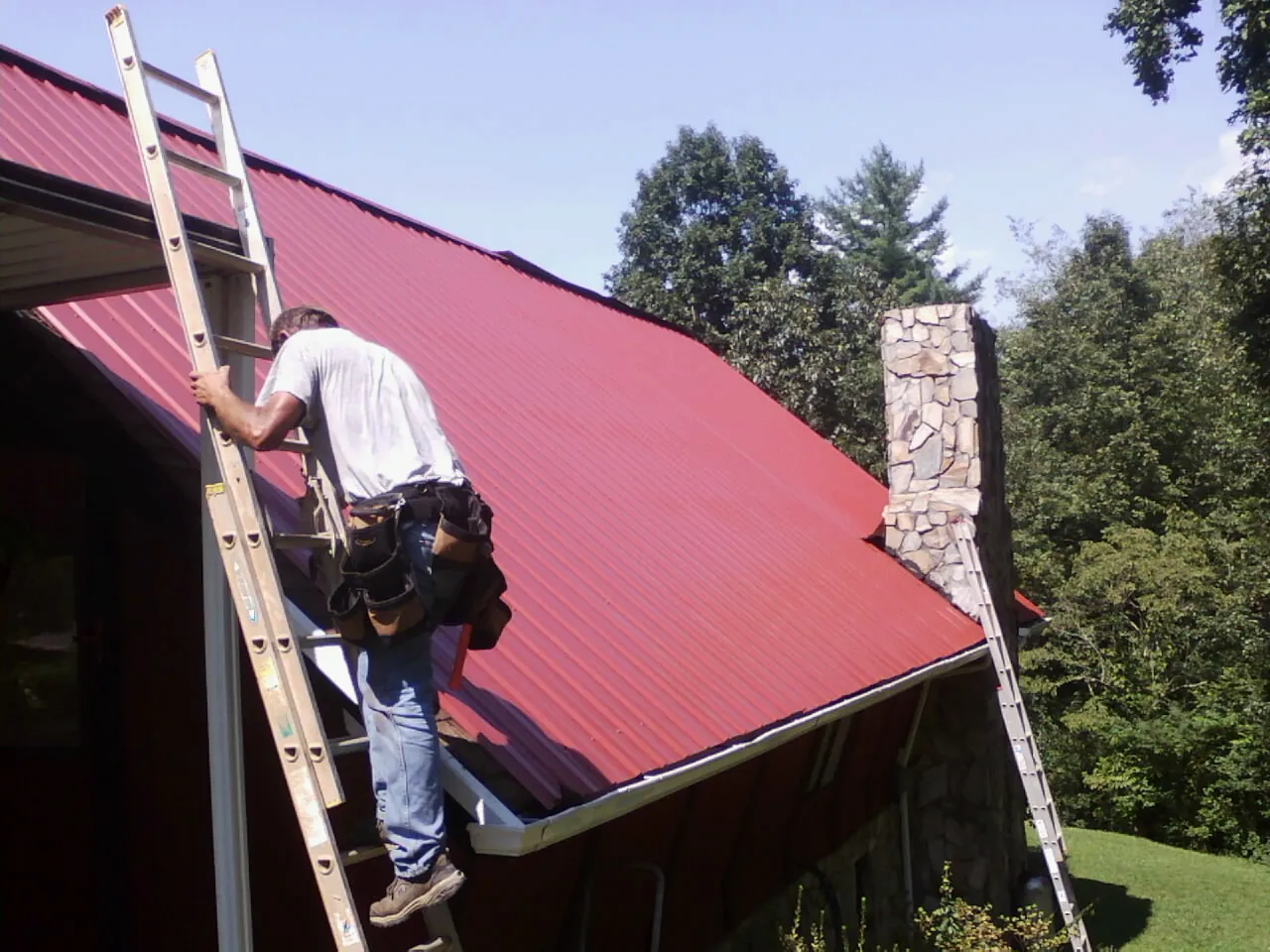 Roofer on a ladder beside a red metal roof and stone chimney — Soffit & Fascia Repair by Roofing Brothers in Butner